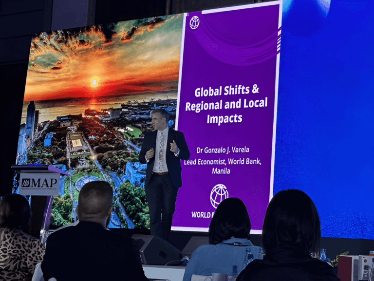 A conference speaker is presenting on the topic 'Global Shifts & Regional and Local Impacts' at the Management Association of the Philippines event. The slide behind the speaker features the World Bank logo and the Manila cityscape during sunset.