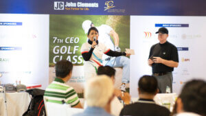 A woman speaking at the John Clements golf tournament at Manila Golf Club, standing next to a man in a black cap. The event banner with sponsor logos is visible in the background.