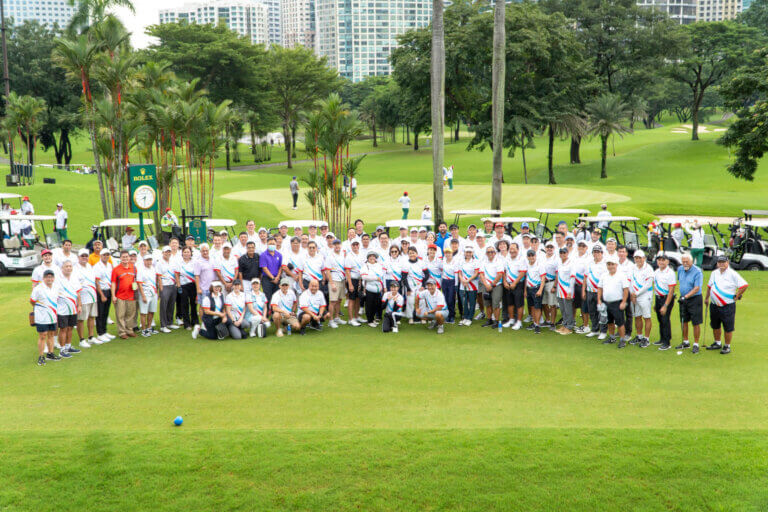 Participants of the John Clements golf tournament at the Manila Golf Club, posing on the green with golf carts and a Rolex clock in the background.