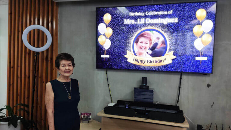 Mrs. Lil Dominguez standing next to a screen displaying a birthday message, with balloons and celebration decorations.