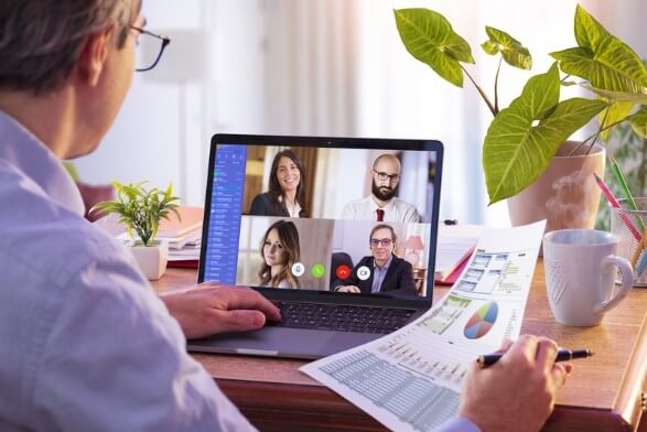 A professional participating in a video conference on a laptop, with four colleagues visible on the screen. The workspace includes charts, a plant, and a coffee mug, symbolizing a remote work setting. This scene illustrates the collaborative nature of remote work recruitment.