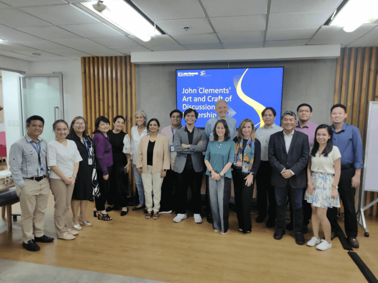 Group photo from the John Clements Art and Craft of Discussion Leadership workshop, standing together in a room. A screen in the background displays the workshop title.