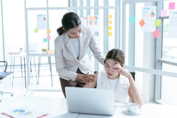 A woman standing beside a seated colleague, offering support by placing a hand on her shoulder. The seated woman appears stressed while working on a laptop, symbolizing empathy in leadership development within the workplace. Colorful sticky notes and charts are visible in the background.