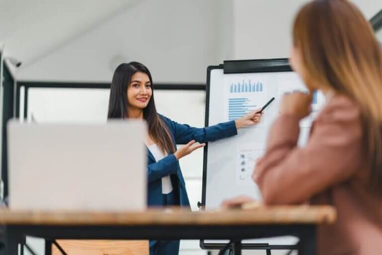 A woman presenting essential leadership skills during a business meeting, pointing to graphs and charts on a flip chart while another person listens attentively.