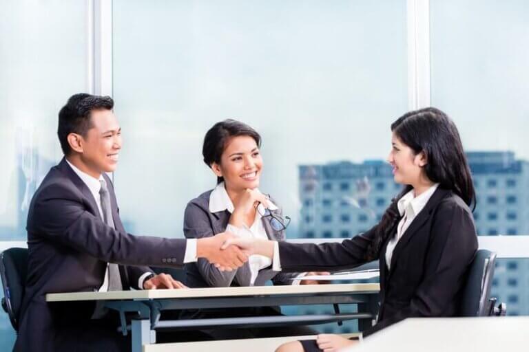 A professional business meeting where two individuals are shaking hands across a desk, while a third person observes and smiles, symbolizing successful collaboration. This scene is representative of the services provided by executive search firms in connecting top talent with leadership roles.