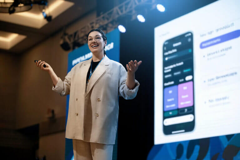 A confident speaker with glasses and a beige suit delivers a presentation on stage. The speaker holds a microphone while gesturing with their hands, engaging the audience with a large screen behind them displaying a mobile interface. This image highlights effective presentation skills through the speaker's clear body language and enthusiasm.