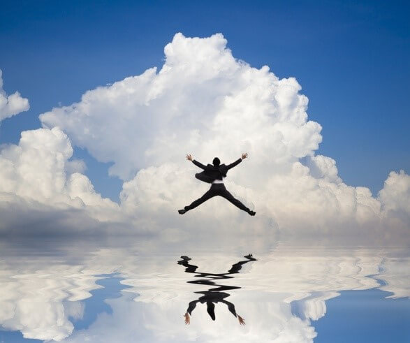 A person in a business suit jumps energetically with arms outstretched against a background of bright clouds and a reflective water surface, symbolizing optimism and career reflection.