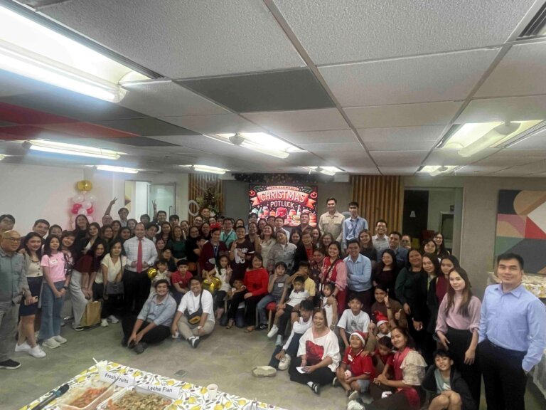 A large group of people gathered in a festive setting for a Christmas potluck party, smiling and posing together with holiday decorations and a food table in the foreground.