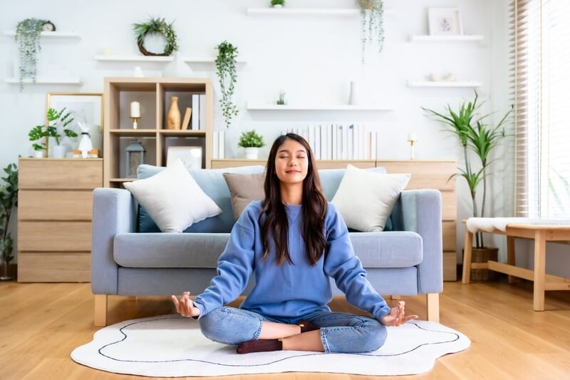 Young woman practicing meditation for emotional healing in a serene living room setting, sitting cross-legged on a rug with a calm expression.