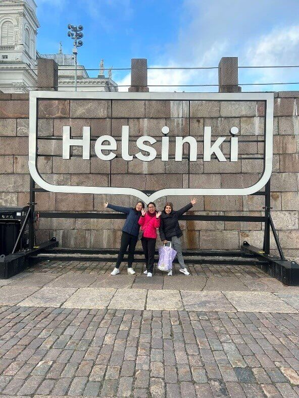 Three smiling individuals pose in front of a large 'Helsinki' sign, symbolizing their involvement in supporting nurses in Finland.