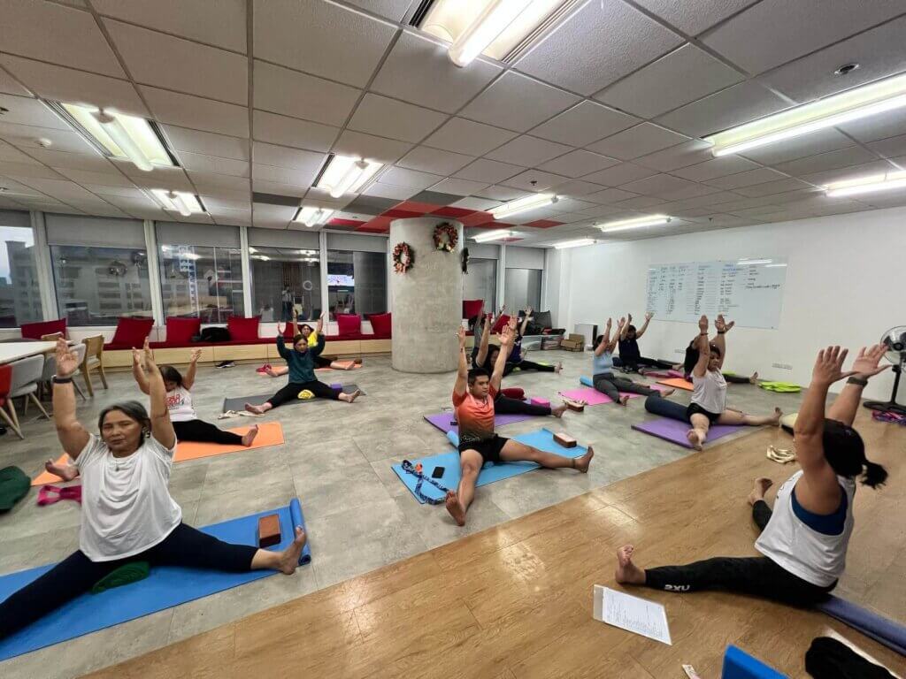 A group of people practicing Iyengar yoga in a well-lit studio, performing seated stretches with arms raised.