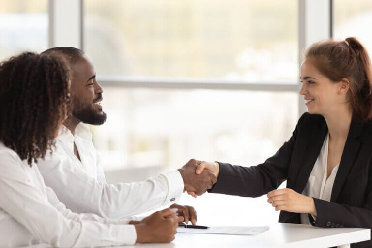 A businesswoman in a black suit shakes hands with a man in a white shirt while another woman sits beside him, symbolizing collaboration and fractional leadership in a professional setting.