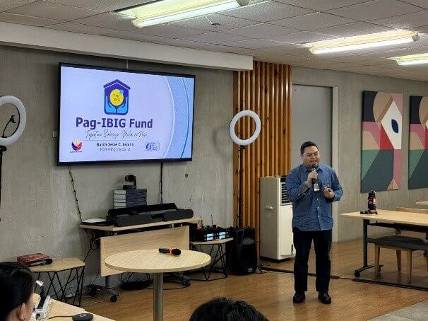 A speaker discusses Pag-IBIG benefits during a presentation at an indoor event, with a screen displaying the Pag-IBIG Fund logo and event details.