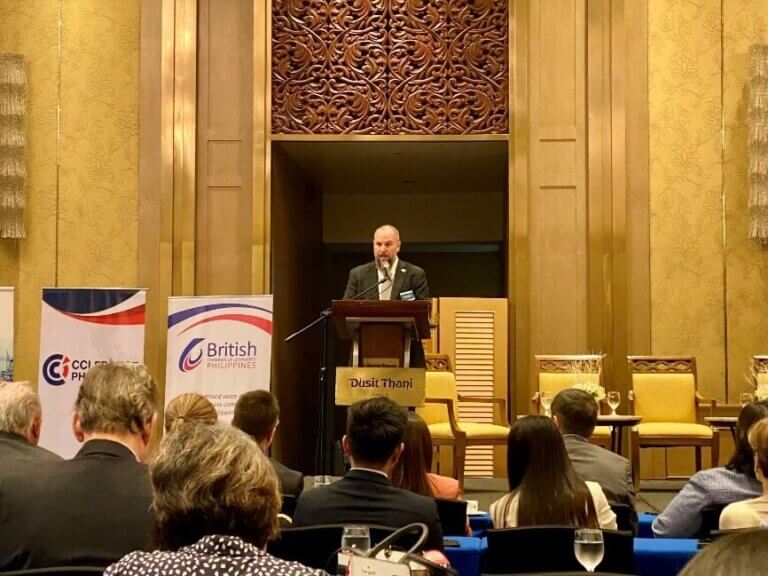 A speaker addresses an audience at a business event discussing investment opportunities in the Philippines, held at Dusit Thani, with banners from British and French chambers of commerce.