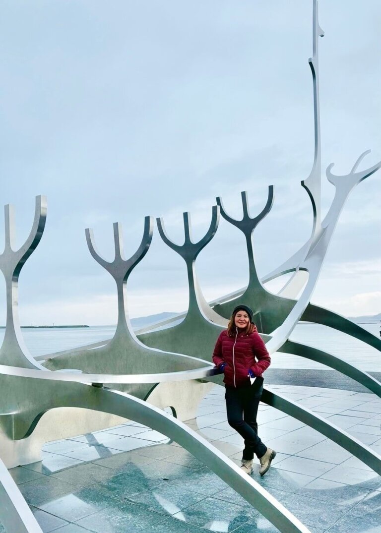 Traveler posing at the Sun Voyager sculpture in Reykjavik, Iceland, with the ocean and distant mountains in the background. A must-visit spot in any Iceland travel guide.