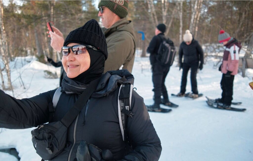 A group of people dressed in winter gear enjoy a snowy adventure in Lapland, with one woman smiling in the foreground while others prepare for snowshoeing in the background.