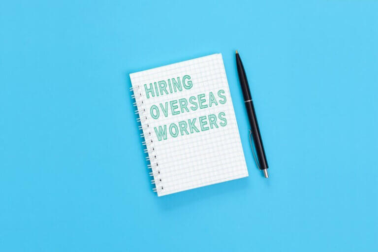 Notebook with 'Hiring Overseas Workers' written on it, representing sectors benefiting from overseas hiring, placed on a blue background with a black pen beside it.