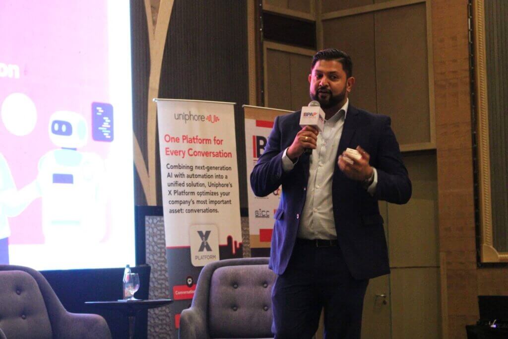 Man in a suit presenting at a conference on AI in customer engagement, with a Uniphore banner highlighting conversational AI solutions in the background.