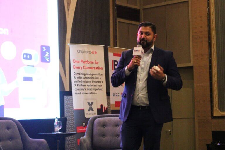 Man in a suit presenting at a conference on AI in customer engagement, with a Uniphore banner highlighting conversational AI solutions in the background.