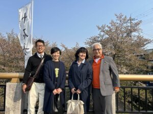 Four professionals stand on a bridge surrounded by cherry blossoms during a business trip to Japan for client engagement. They are dressed in business casual attire, with a white flag bearing Japanese text visible on the left side, adding cultural context to the scene.