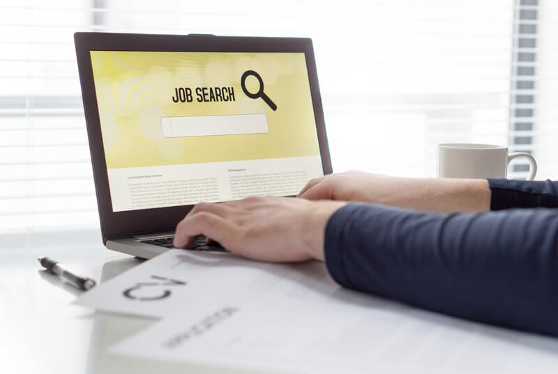 Person using a laptop with a "Job Search" screen and a printed CV nearby, symbolizing efforts to find jobs.