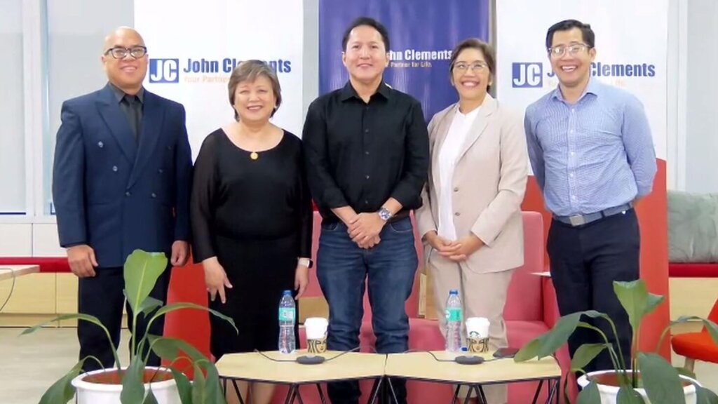 A diverse group of five professionals stands in front of a backdrop featuring the text 'John Clements,' dressed in business attire. The setting, complete with potted plants and a table with water bottles and coffee cups, represents integrity in leadership in the Philippines, highlighting professionalism and ethical leadership in corporate or HR environments.