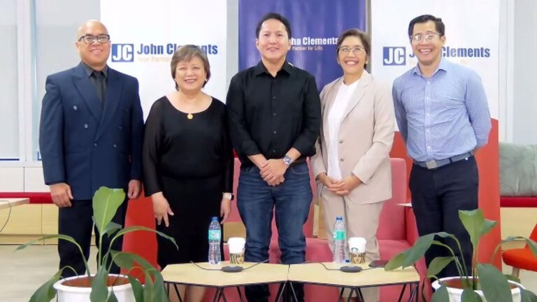A diverse group of five professionals stands in front of a backdrop featuring the text 'John Clements,' dressed in business attire. The setting, complete with potted plants and a table with water bottles and coffee cups, represents integrity in leadership in the Philippines, highlighting professionalism and ethical leadership in corporate or HR environments.