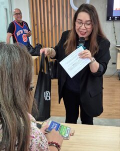A woman presents on nontraditional career paths in the Philippines, holding a microphone and a paper while handing a black bag to a seated participant. The setting suggests a seminar or workshop, highlighting alternative career opportunities.