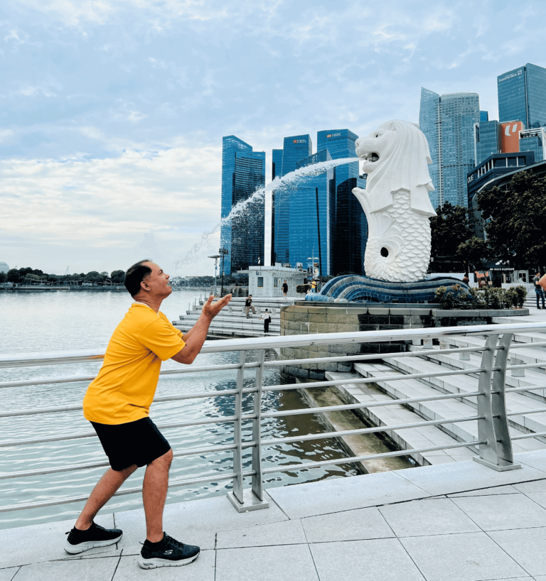 A traveler in a yellow shirt poses playfully near the Merlion statue, catching its water spray, with Singapore’s skyline in the background. A snapshot of fun during a Singapore family travel adventure.