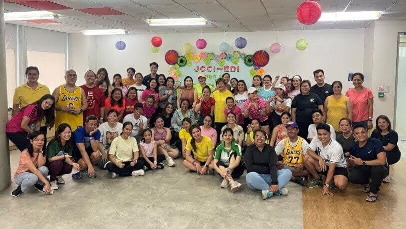 Group photo of employees smiling and wearing colorful outfits during one of John Clements’ workplace wellness events, with festive decorations in the background.